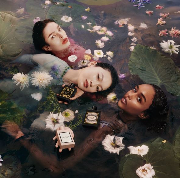 Three women lying in water with lily pads and flowers, holding perfume bottles.