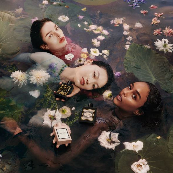 Three women lying in water with lily pads and flowers, holding perfume bottles.