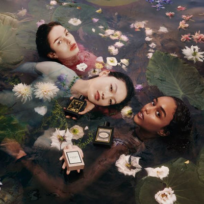 Three women lying in water with lily pads and flowers, holding perfume bottles.