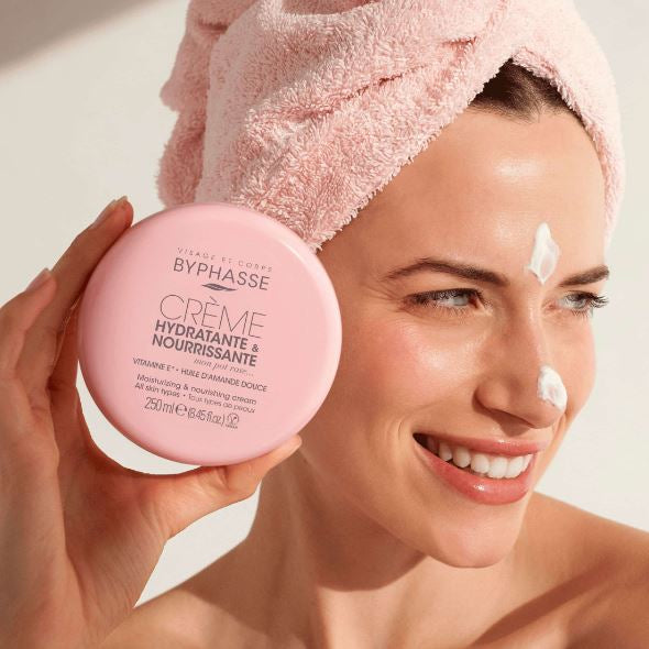Woman applying Byphasse cream to her face with a pink towel on her head.