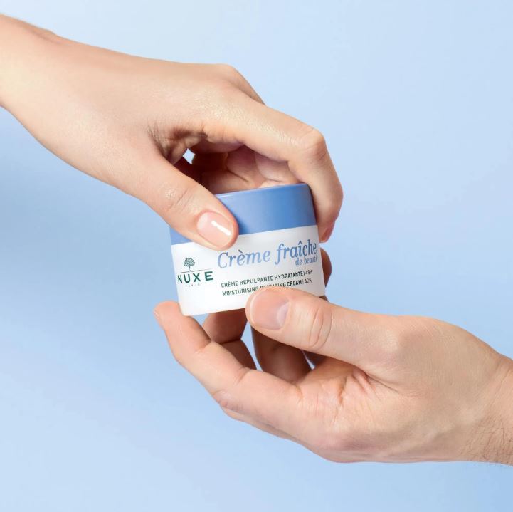 Hand holding a Nuxe Crème fraçìhe jar against a light blue background