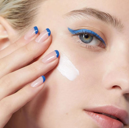 Close-up of a woman's face with blue eyeliner and matching nail polish, applying cream to her cheek.