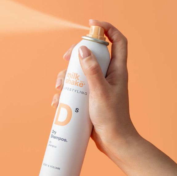 Hand holding a bottle of Milk Shake Dry Shampoo against an orange background
