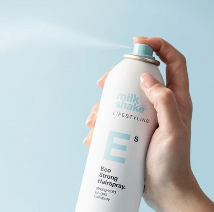 Hand holding a can of Milk Shake Eco Strong Hairspray against a light blue background