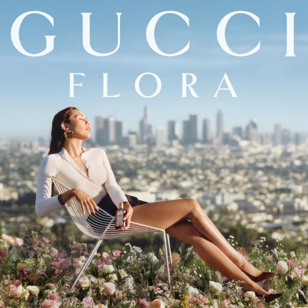 Woman sitting on a chair in a field of flowers with a cityscape in the background, Gucci Flora branding.