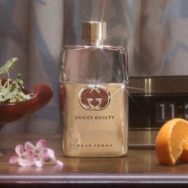 Gucci Guilty perfume bottle on a table with flowers and an orange.