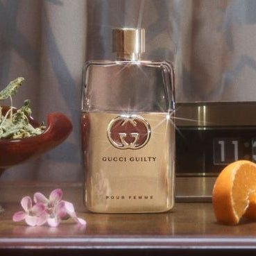 Gucci Guilty perfume bottle on a table with flowers and an orange.