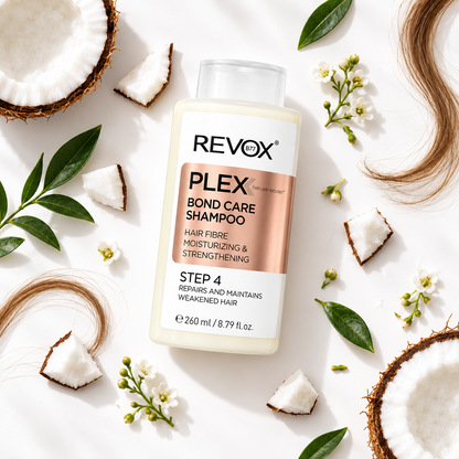 A bottle of REVOX PLEX Bond Care Shampoo Step 4 is displayed in a styled flat-lay arrangement on a white background, surrounded by coconut halves, green leaves, small white flowers, and strands of brown hair.