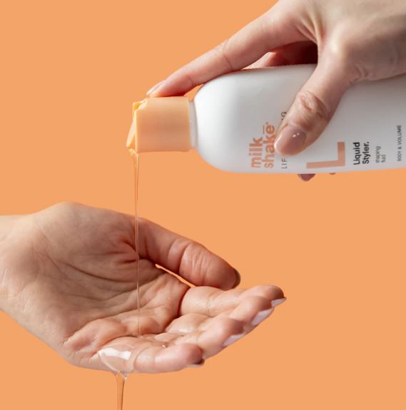 Hand holding a bottle of 'milk shake' liquid styling product with orange background