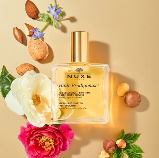 Nuxe Huile Prodigieuse bottle surrounded by flowers and nuts on a beige background