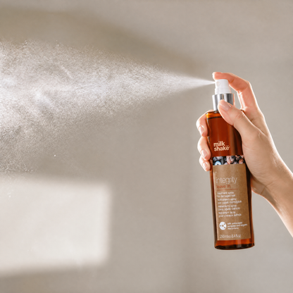 Hand holding a 'milk_shake' hair spray bottle and spraying it against a neutral background