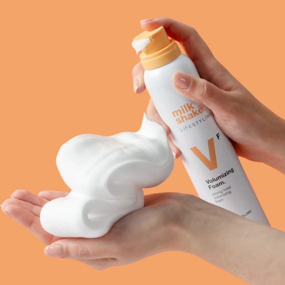 Hand holding a bottle of 'milk_shake' Volumizing Foam against an orange background